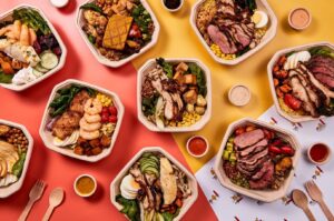 A variety of healthy restaurant foods displayed in plastic containers, showcasing colorful salads, grains, and proteins.