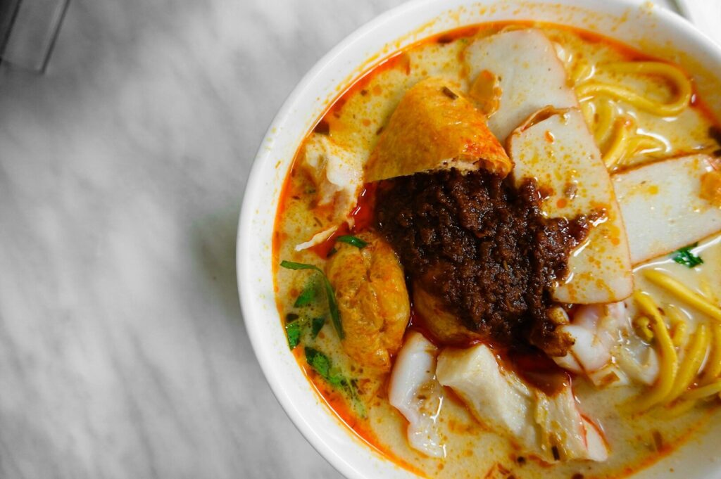 A steaming bowl of laksa featuring noodles and tender meat, surrounded by a rich, flavorful broth.
