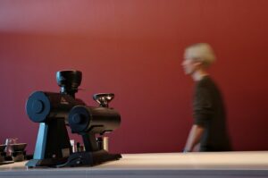 A blurred person walks past black coffee grinders on a countertop, set against a deep red wall, creating a minimalist, modern cafe ambiance.