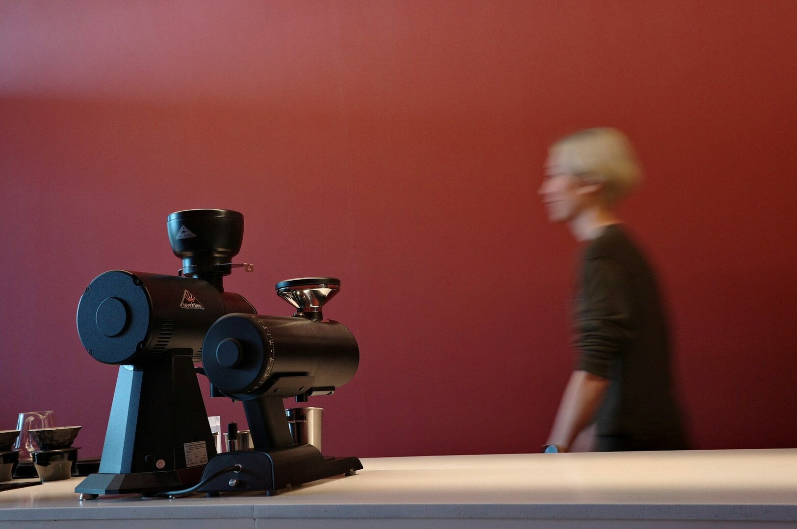 A blurred person walks past black coffee grinders on a countertop, set against a deep red wall, creating a minimalist, modern cafe ambiance.