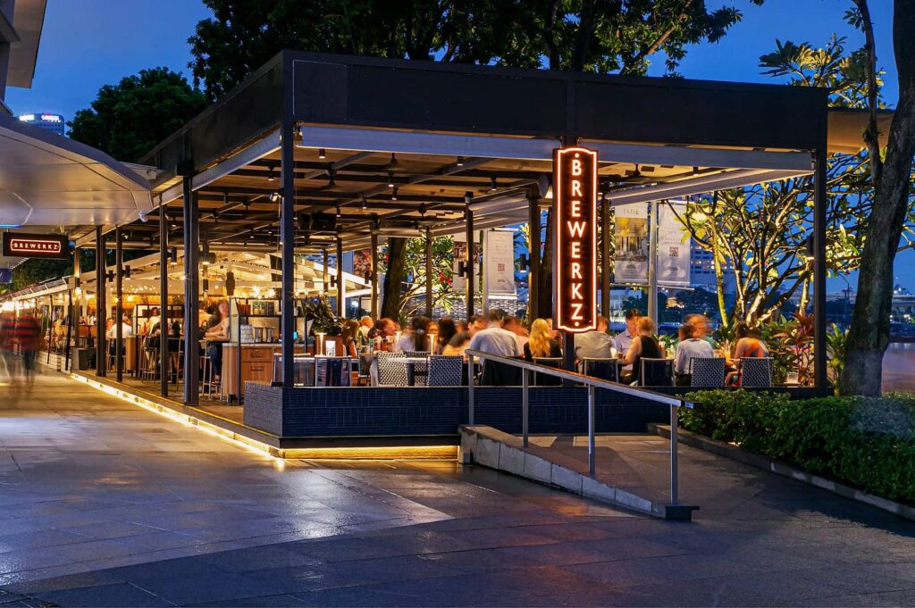 Outdoor evening scene of a lively restaurant with a glowing "Brewerkz" sign. Diners enjoy meals under warm lights, surrounded by greenery and a riverside view.