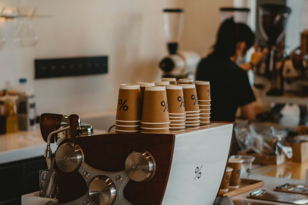 A stack of paper coffee cups with a percent sign logo sits on a modern espresso machine. A barista works in the background, creating a busy café ambiance.
