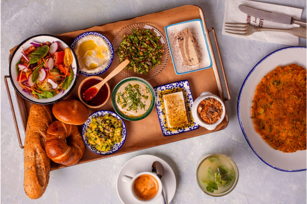 A brunch spread featuring a tray with salad, hummus, bread, dips, and grains. Beside it are a cappuccino, a refreshing drink, and a large flatbread.