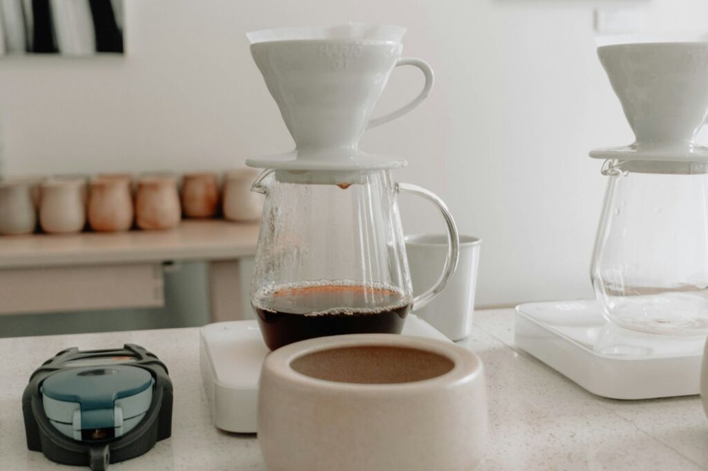 White ceramic pour-over coffee makers brewing into glass carafes on a countertop, with a relaxed and minimalist café setting in warm tones.