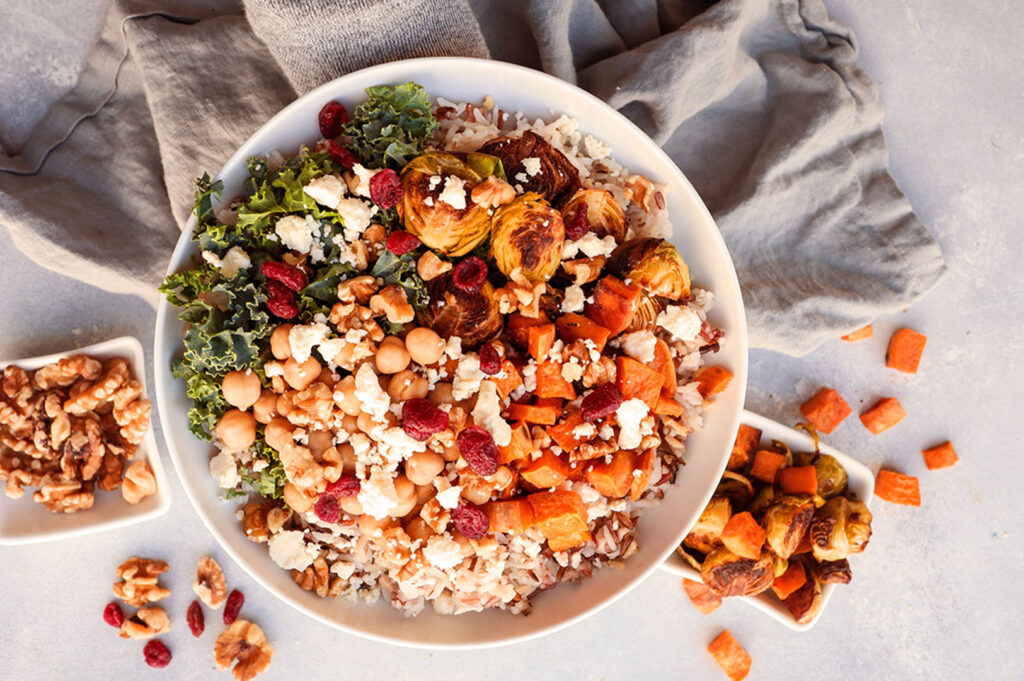 A colorful salad bowl with chickpeas, kale, roasted Brussels sprouts, sweet potatoes, cranberries, walnuts, and feta on a gray cloth. Nut and veggie dishes nearby.