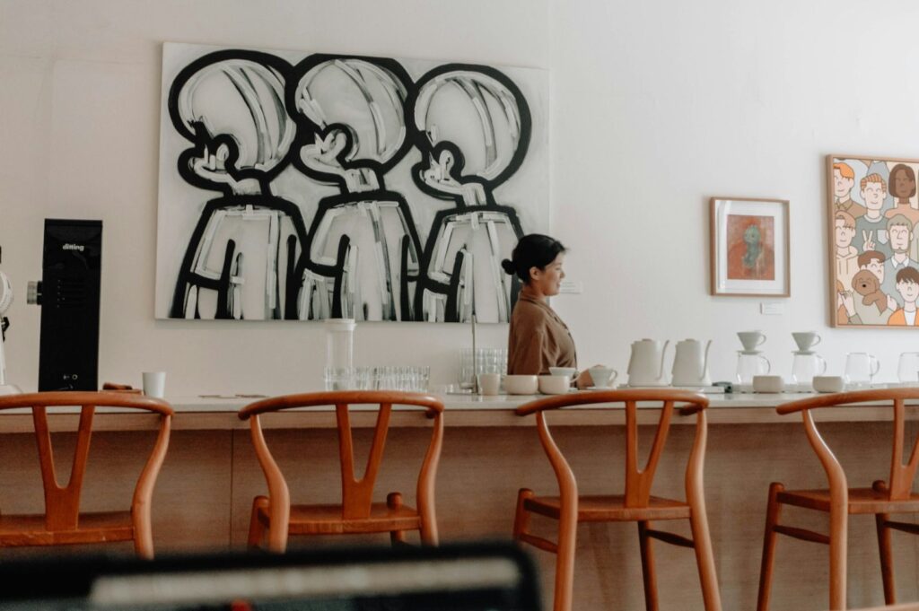 A woman stands at a counter in a minimalist café, with wooden chairs along the bar. Bold black-and-white art and colorful portraits adorn white walls.