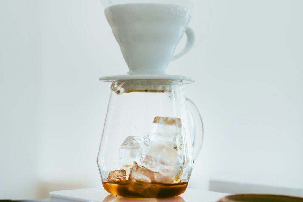 A white ceramic coffee dripper sits atop a clear glass carafe, brewing over ice cubes. The scene is bright and minimalistic, conveying freshness.