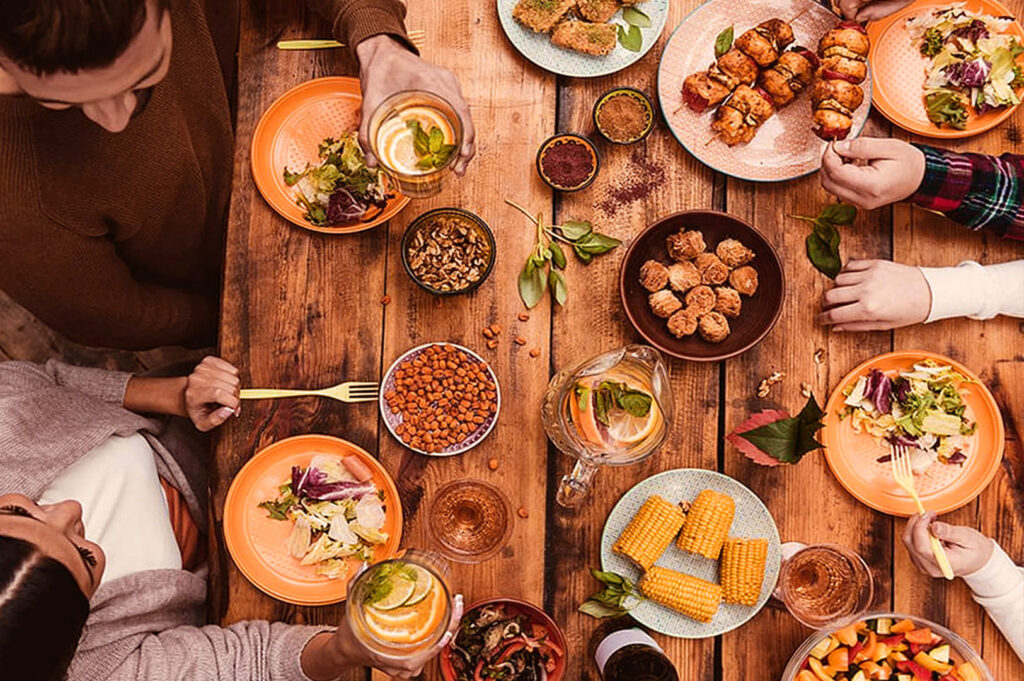 Top view of a wooden table filled with colorful plates of food, including salad, corn, kebabs, and bowls of nuts. Hands are reaching for dishes, creating a warm, communal atmosphere.