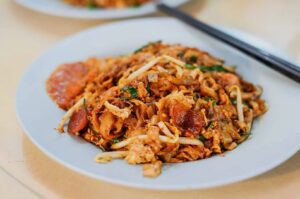 A serving of char kway teow on a plate, accompanied by chopsticks and a fork, highlighting its rich textures and colors.