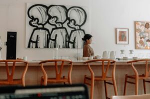 A woman stands in a cafe with wooden chairs and a counter in the foreground. Abstract art of three figures and colorful portraits decorate the walls.