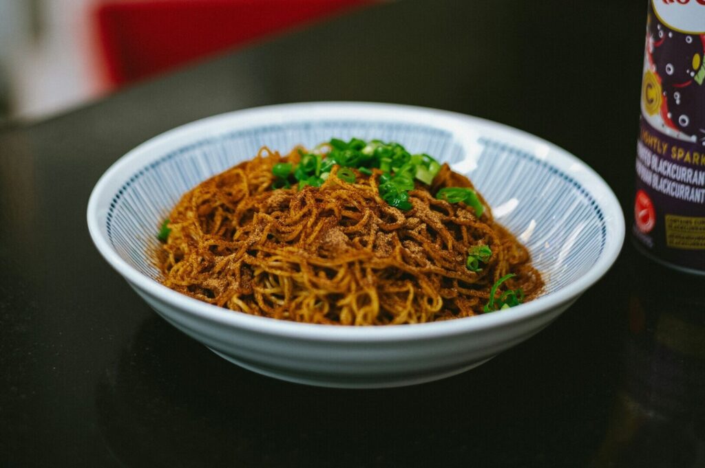 A bowl filled with Singapore noodles accompanied by a bottle of sauce, ready for a flavorful dining experience.
