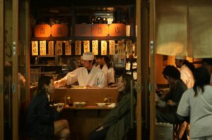 Cozy Japanese sushi bar with chefs preparing food behind the counter. Patrons sit and chat, enjoying a warm, welcoming atmosphere. Lanterns and calligraphy hang above.