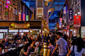 Bustling night market with people dining outdoors. Vibrant signs and food stalls line the street, creating a lively, colorful atmosphere.