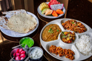 A vibrant Indian vegetarian thali on a table with rice, curries, dal, and fried snacks. Accompanied by roti, salad, yogurt, and a colorful fruit platter.