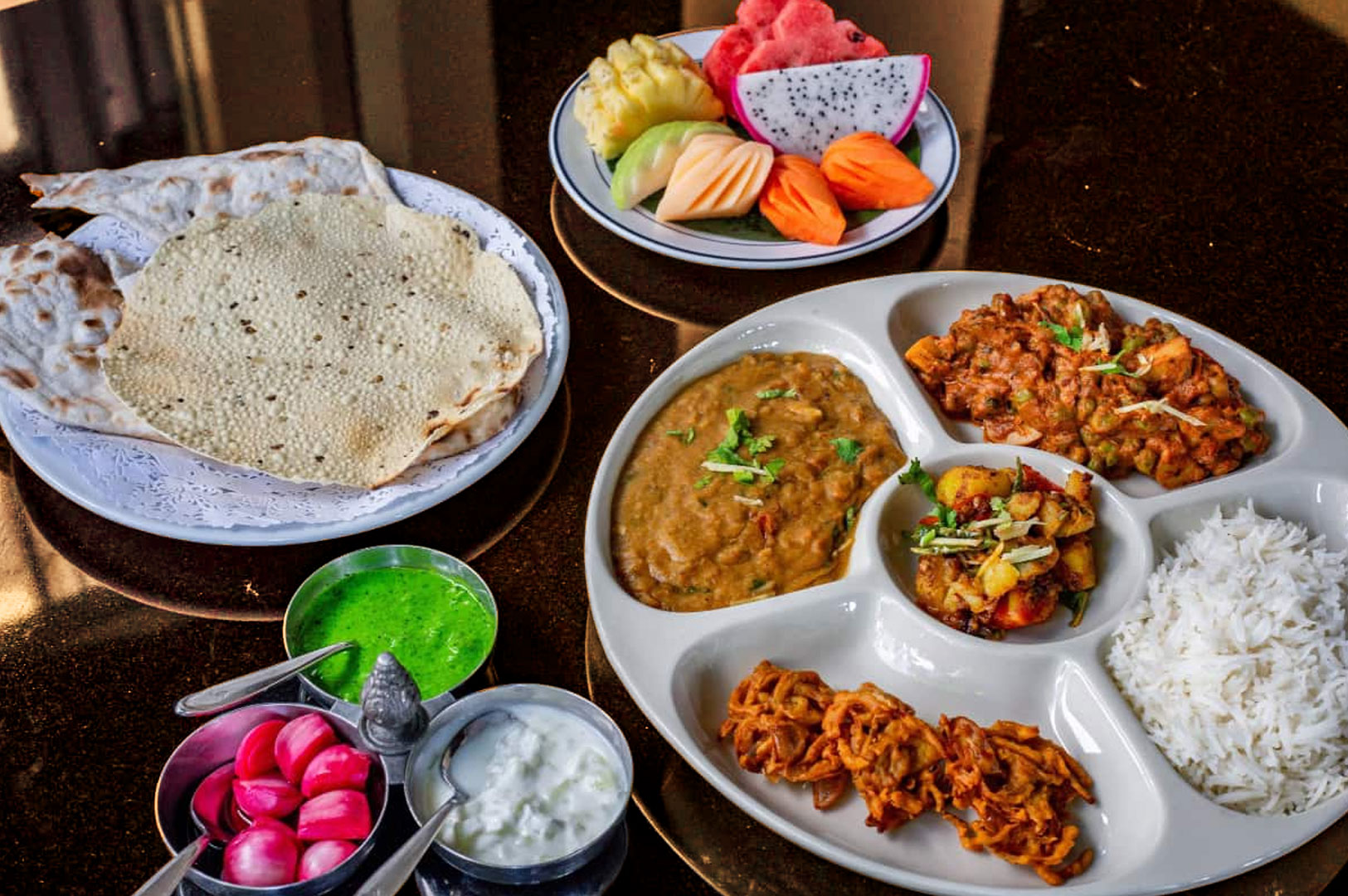 A vibrant Indian vegetarian thali on a table with rice, curries, dal, and fried snacks. Accompanied by roti, salad, yogurt, and a colorful fruit platter.