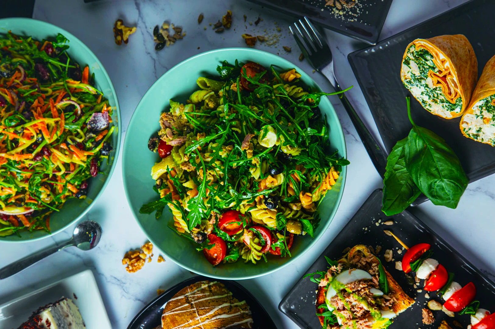 Aerial view of a colorful spread featuring a pasta salad with arugula, tomato, and olives, wraps, bruschetta, cake, and capprese. Fresh and inviting.