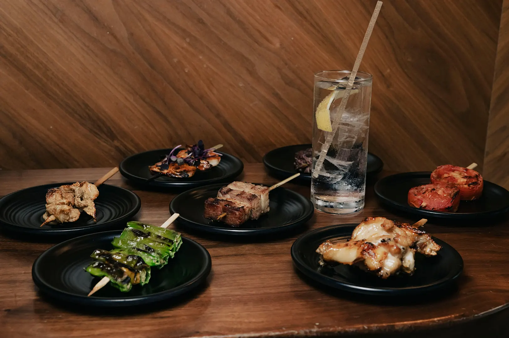 A variety of grilled Japanese yakitori skewers on small black plates with a iced drink, showcasing healthy izakaya dishes on a wooden table.