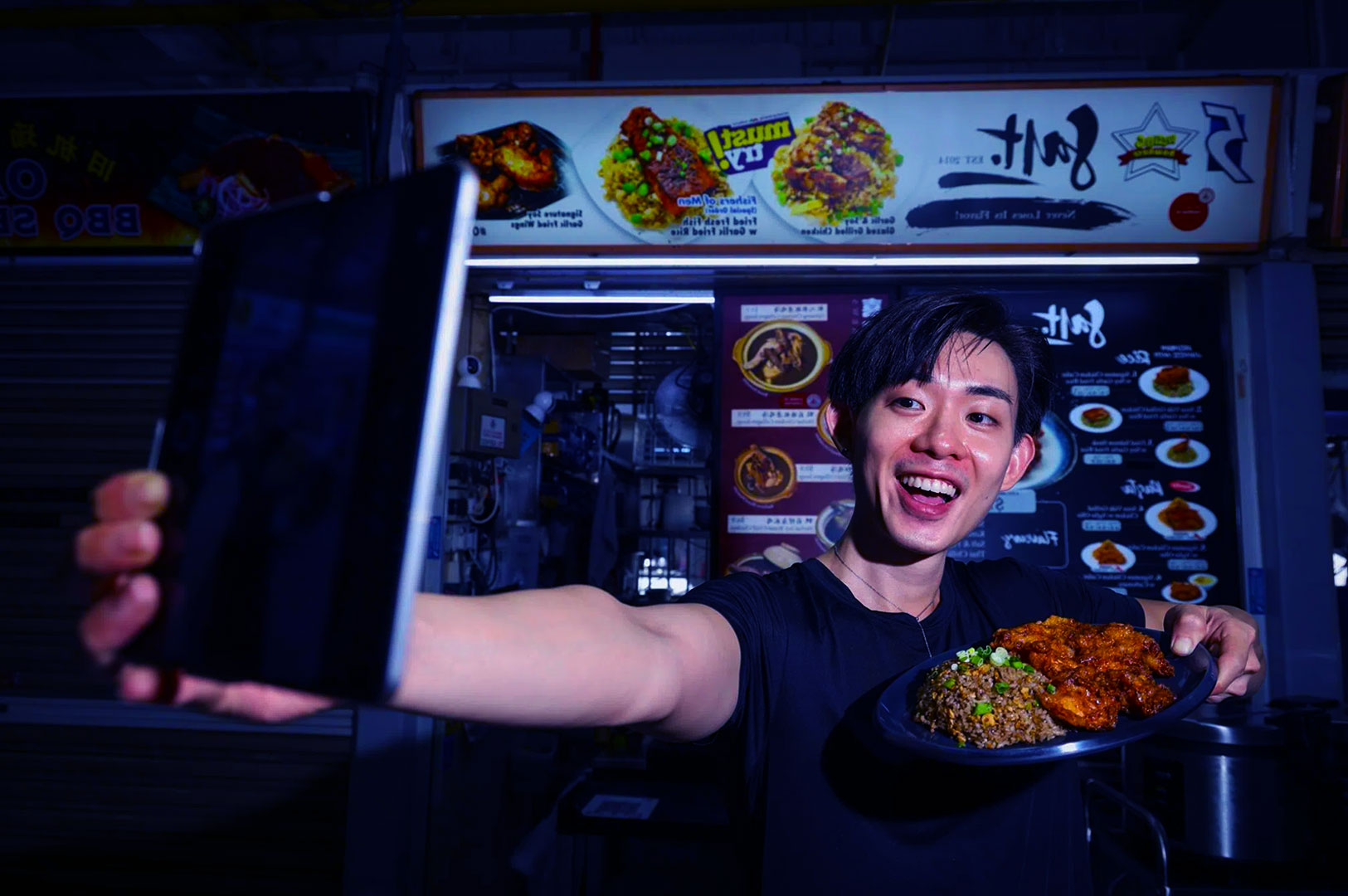 A person smiling and holding a plate of food, taking a selfie at a food stall. Menus and colorful dishes displayed in the vibrant background.