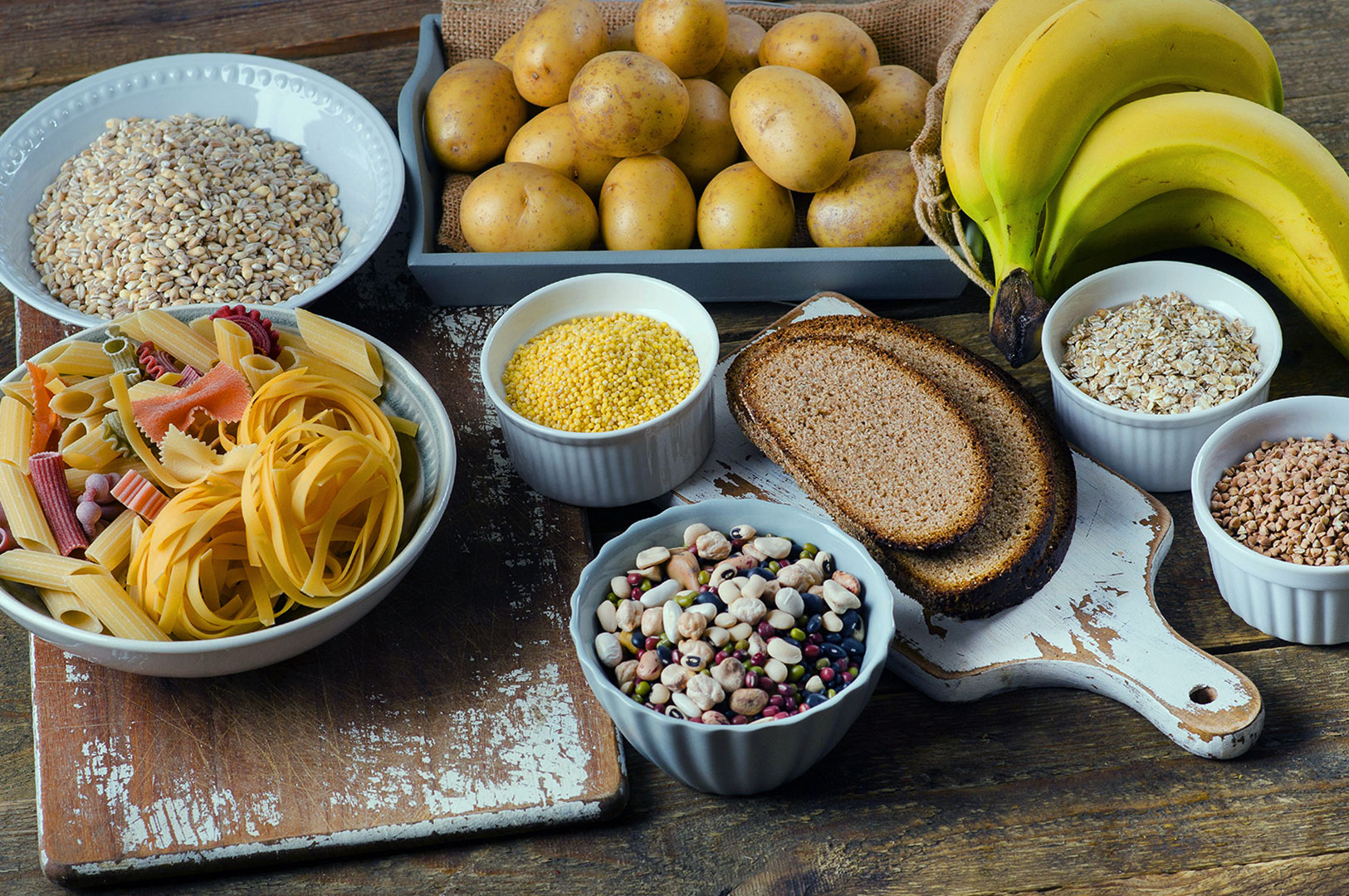 A rustic display of carbohydrates: potatoes, bananas, pasta, grains, beans, bread, and oats arranged on wooden boards, evoking a nourishing, natural feel.