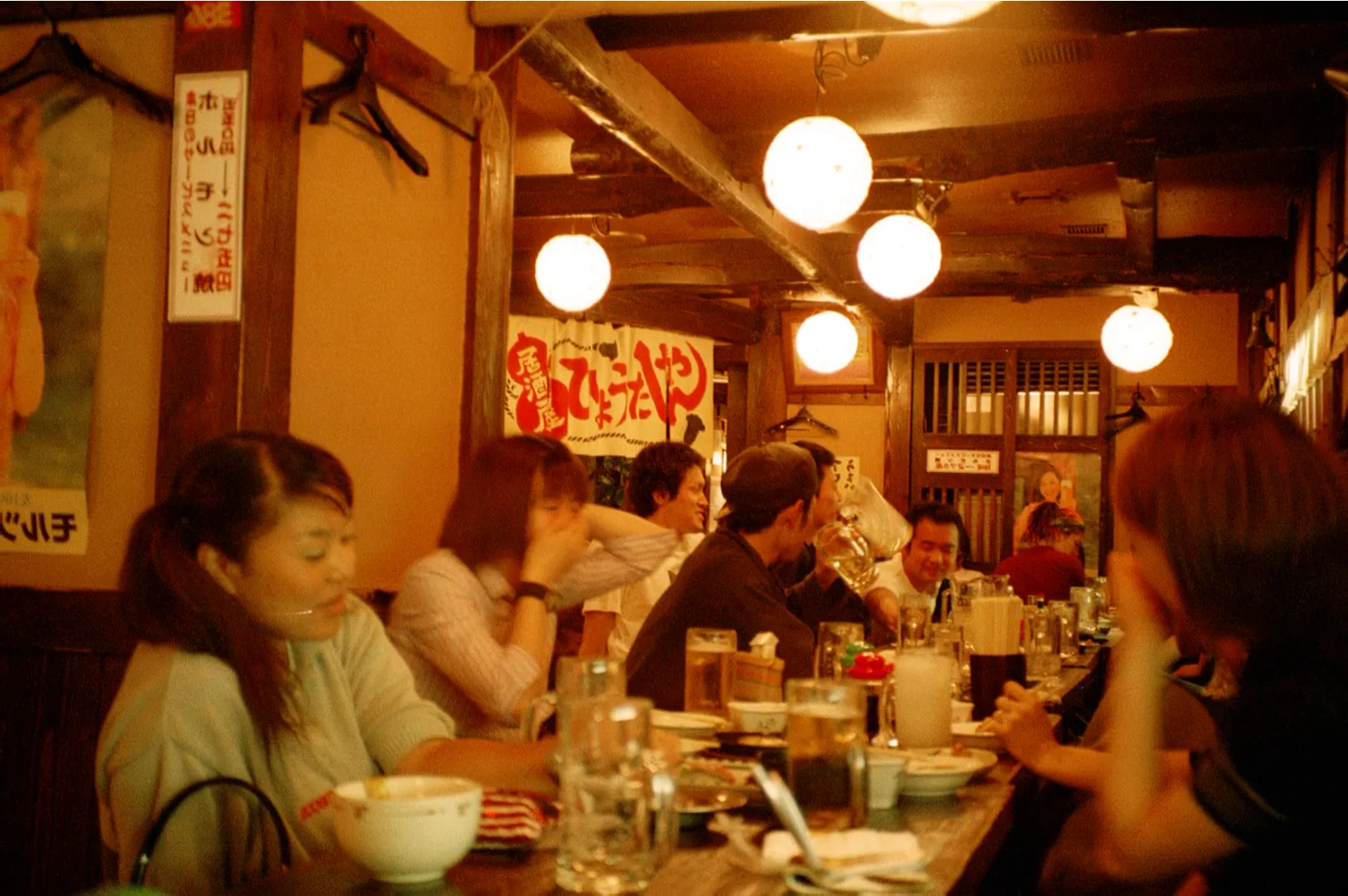 Narrow, bustling alley in Japan, illuminated by warm red paper lanterns and bright signs. People chat outside cozy bars, evoking a lively, inviting atmosphere.