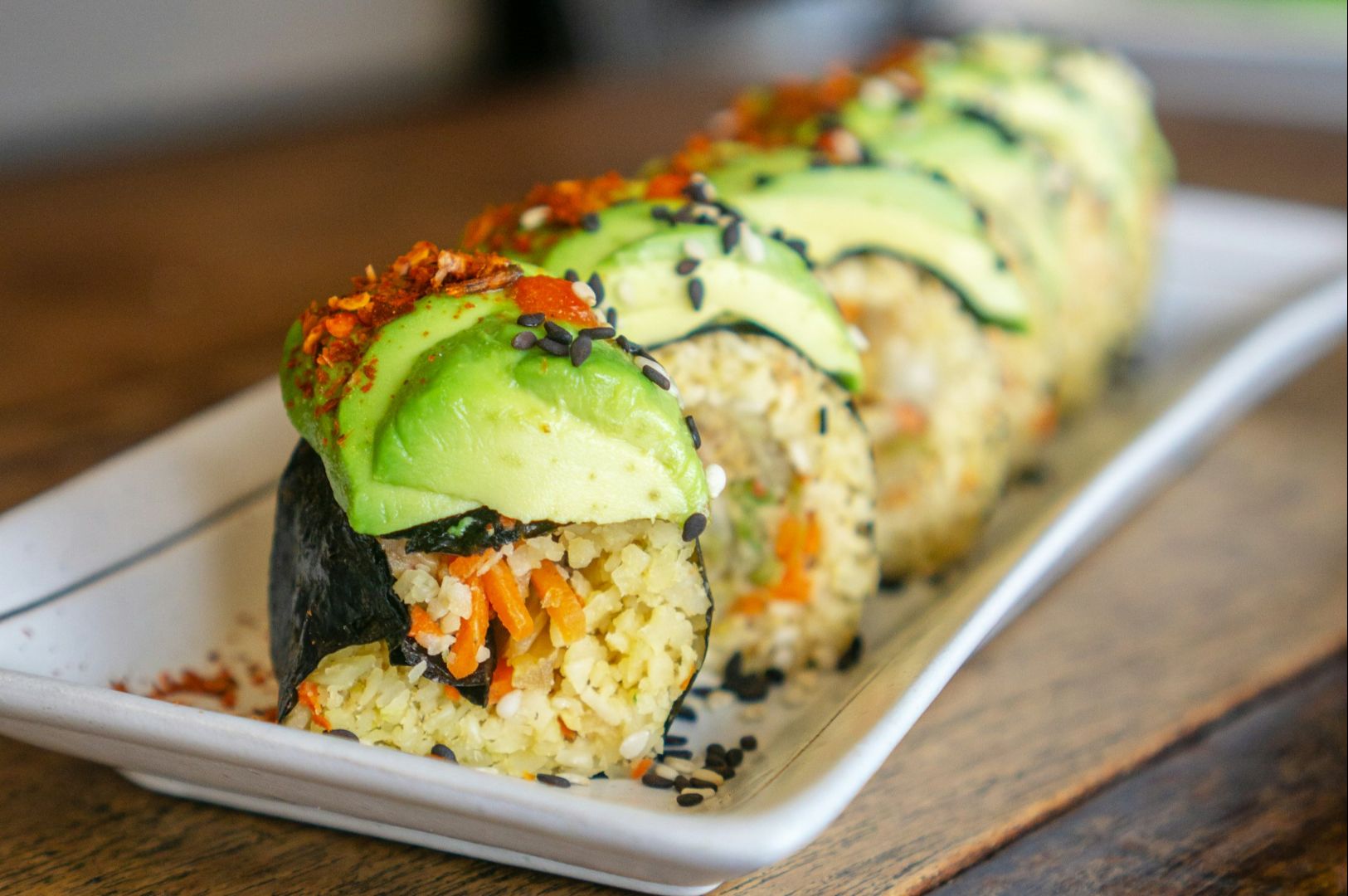 A close-up of a sushi roll on a white plate, topped with avocado slices, sprinkled with black sesame seeds and chili flakes. The roll contains rice and vegetables.
