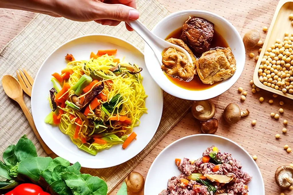 A hand serves tofu from a saucy bowl, next to vibrant noodle and rice dishes with vegetables. Soybeans and mushrooms are arranged around for natural freshness.