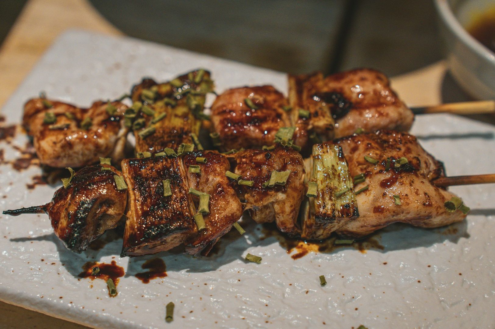 Grilled chicken skewers garnished with herbs, glistening with sauce, served on a textured white plate. The scene suggests warmth and savoriness.