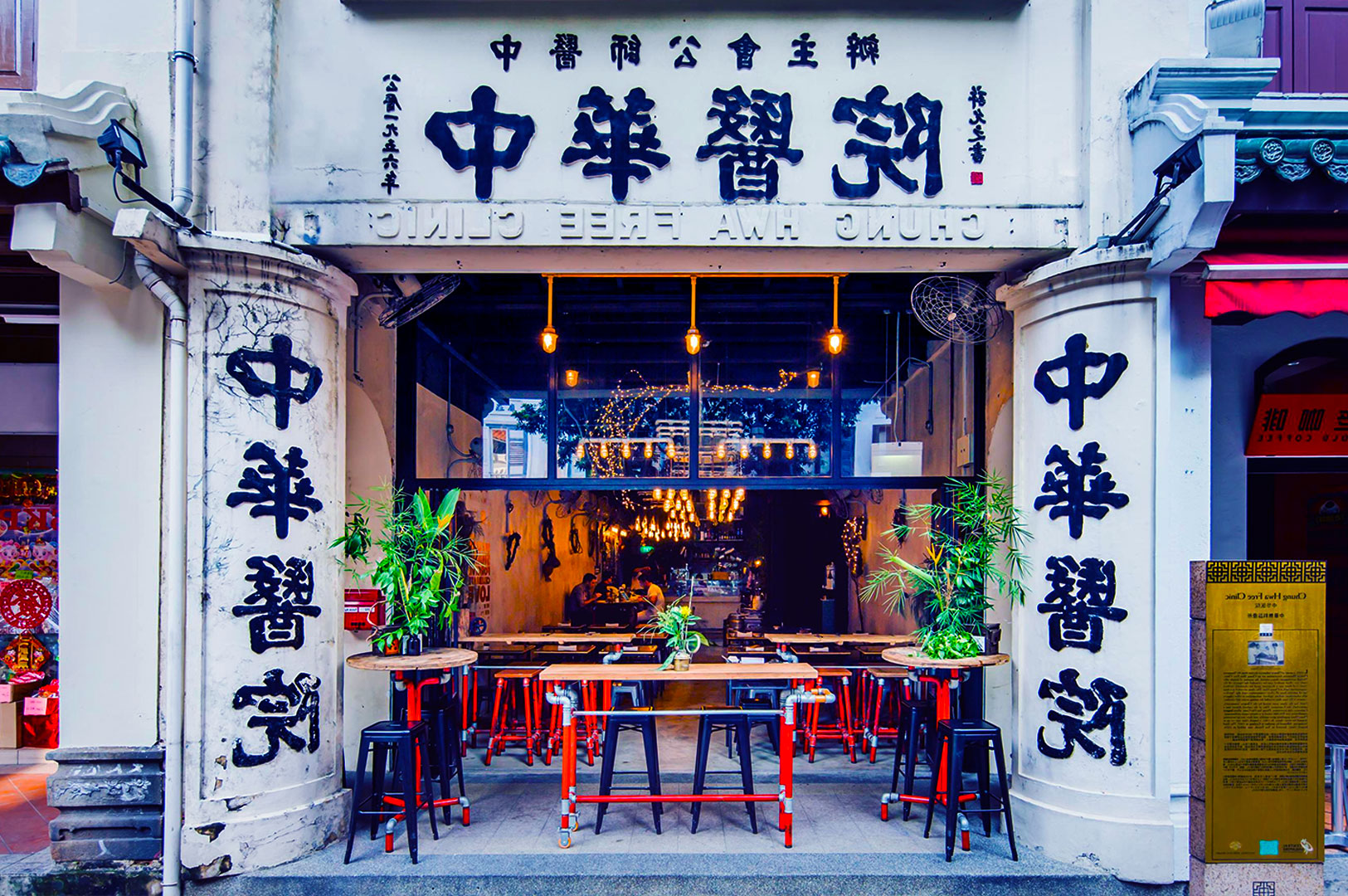 Street view of a Chinese-style cafe with bold black and white signage. The entrance features wooden tables, red stools, and potted plants, creating a welcoming ambiance.