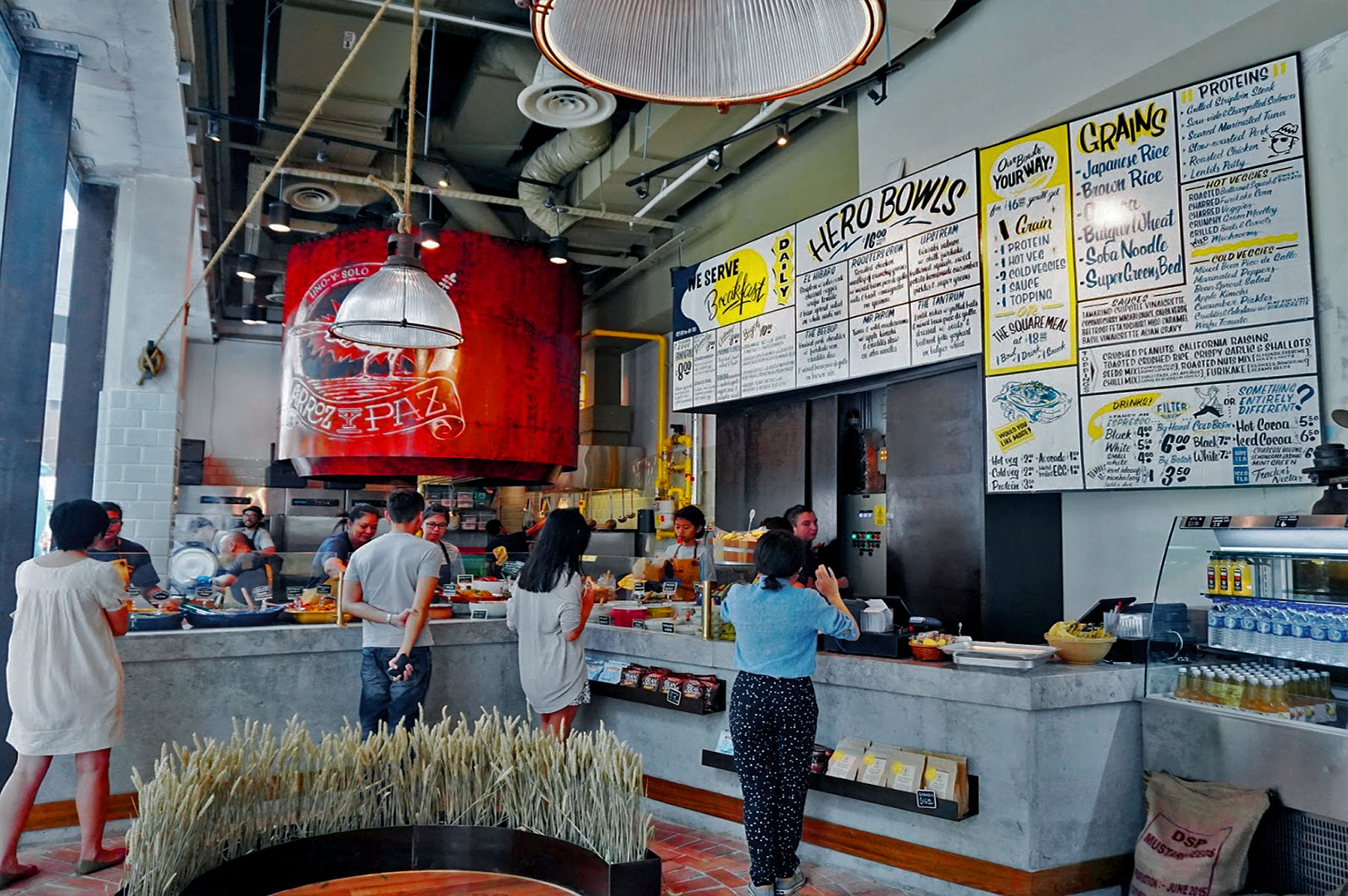 Casual restaurant interior with customers ordering at a counter. Large menu boards display various food options. Industrial decor with a welcoming vibe.