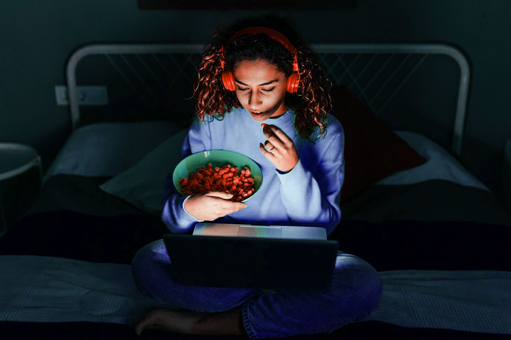 A person sits cross-legged on a bed, wearing headphones, intently watching a laptop screen in a dimly lit room. They hold a bowl of snacks.