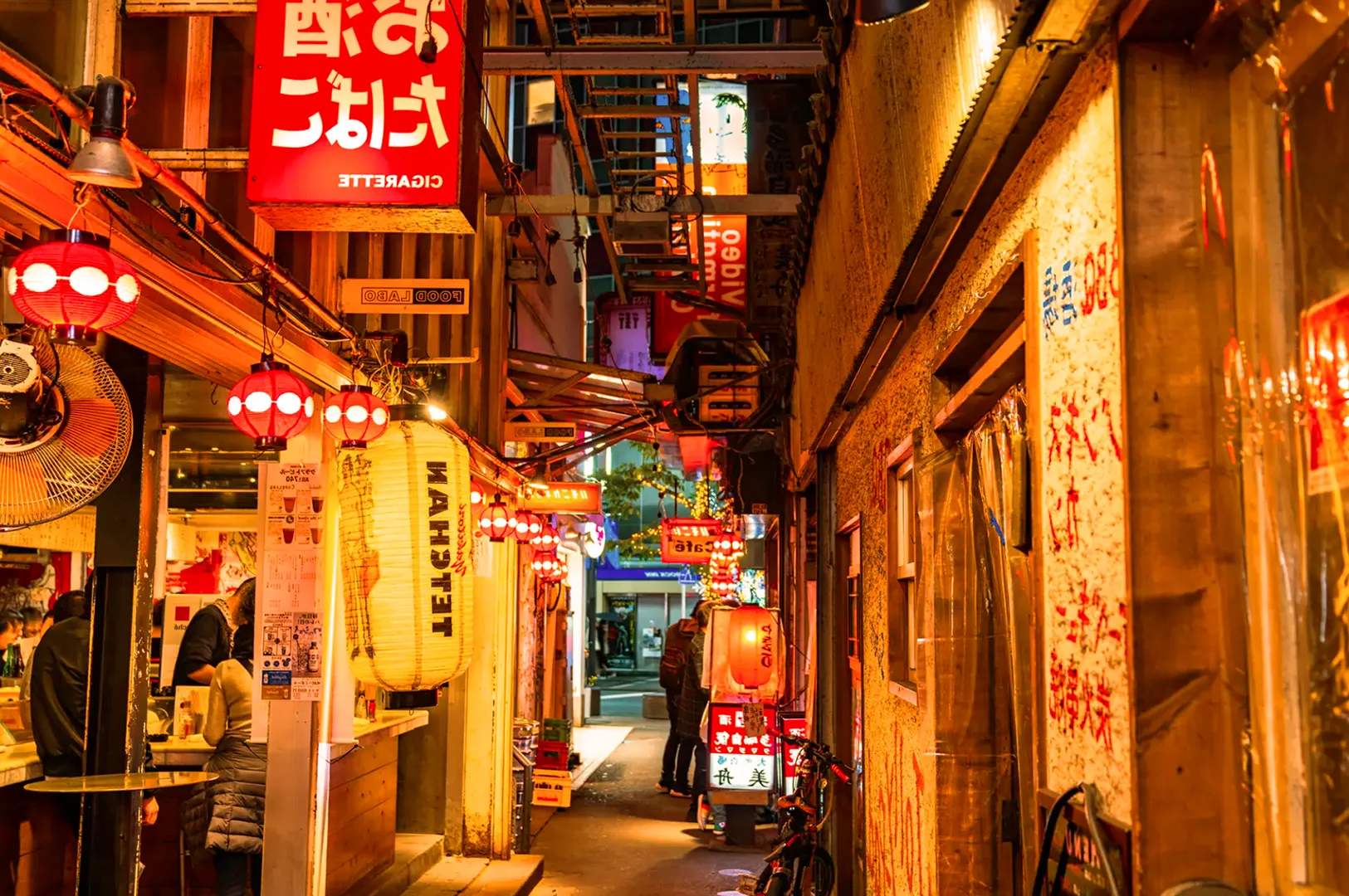 Narrow, bustling alley in Japan, illuminated by warm red paper lanterns and bright signs. People chat outside cozy bars, evoking a lively, inviting atmosphere.