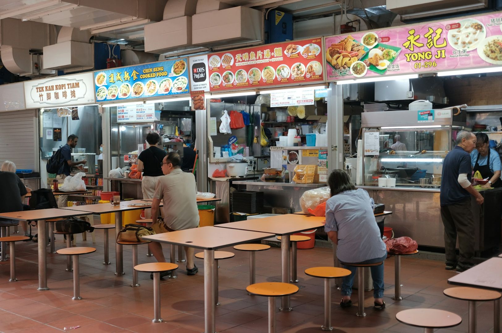 Indoor scene of a lively hawker center with colorful food stalls. People are seated at simple tables, enjoying meals, creating a bustling, communal atmosphere.