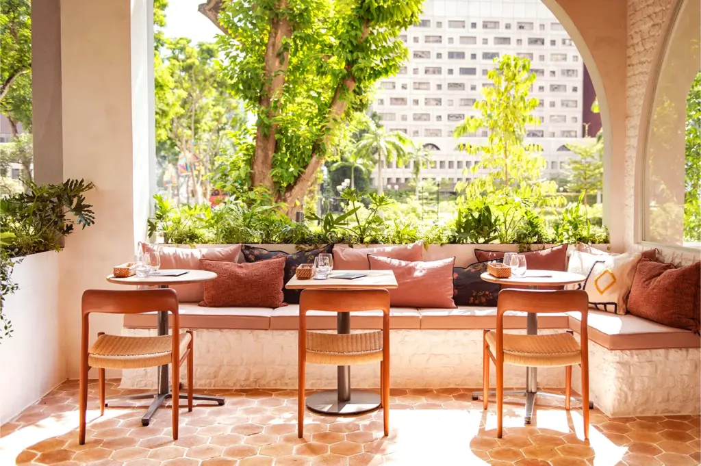 Sunny outdoor café with cozy seating, wooden chairs, and round tables. Greenery surrounds the area, providing a serene, inviting ambiance.