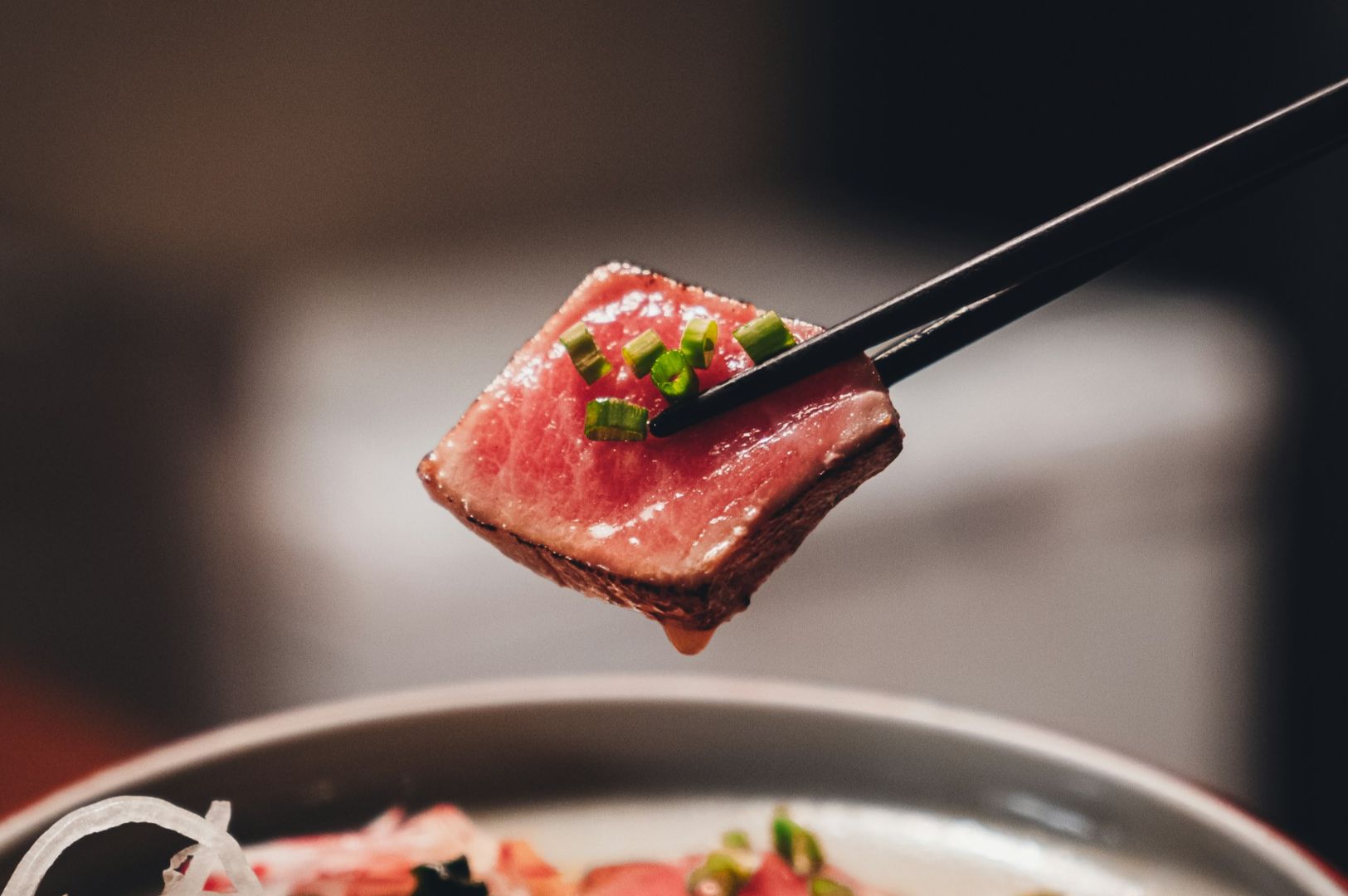 Close-up of chopsticks holding a seared tuna slice garnished with green onions above a dish. The ambiance is warm and appetizing.