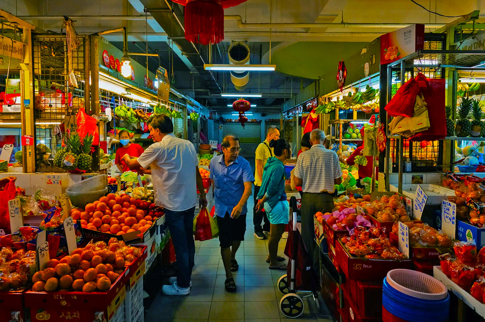Bustling market scene with people shopping for fruits and vegetables under warm lights. Stalls display bright produce, creating a lively, vibrant atmosphere.