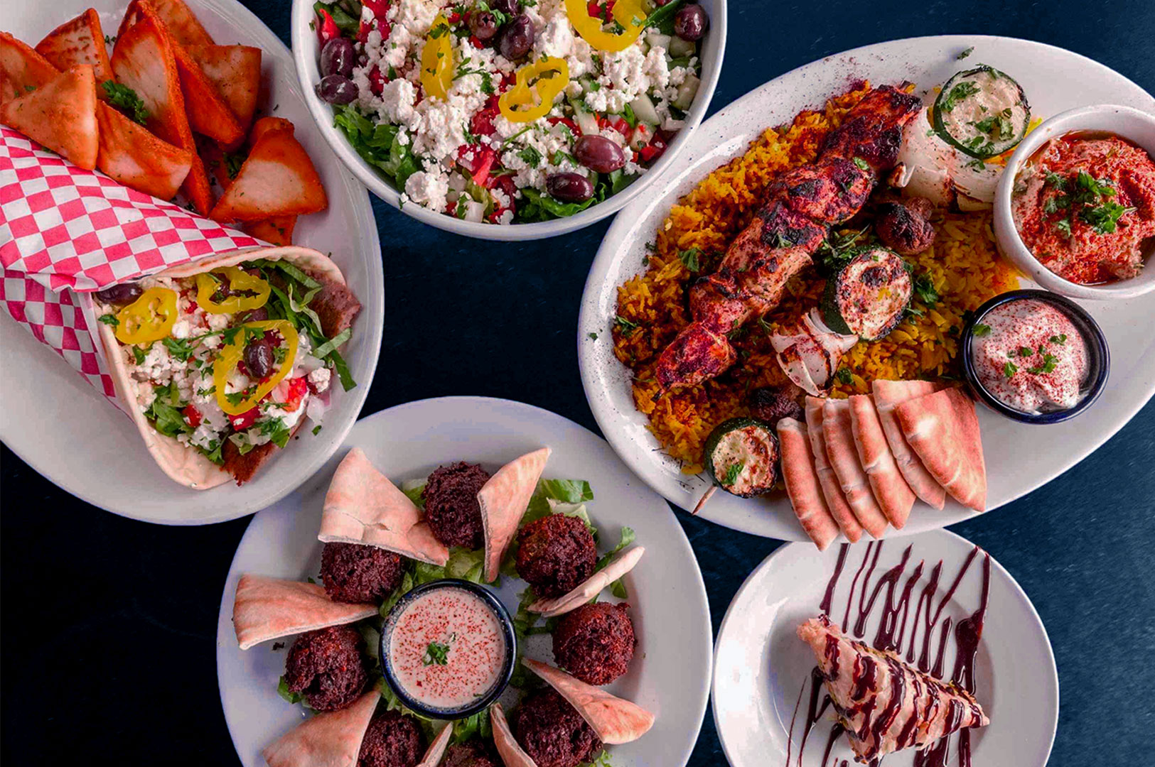 An assortment of Greek dishes is artfully arranged on a dark background. Plates feature gyros, salad, skewers, dips, pita bread, and a dessert slice, evoking a vibrant and savory feast.