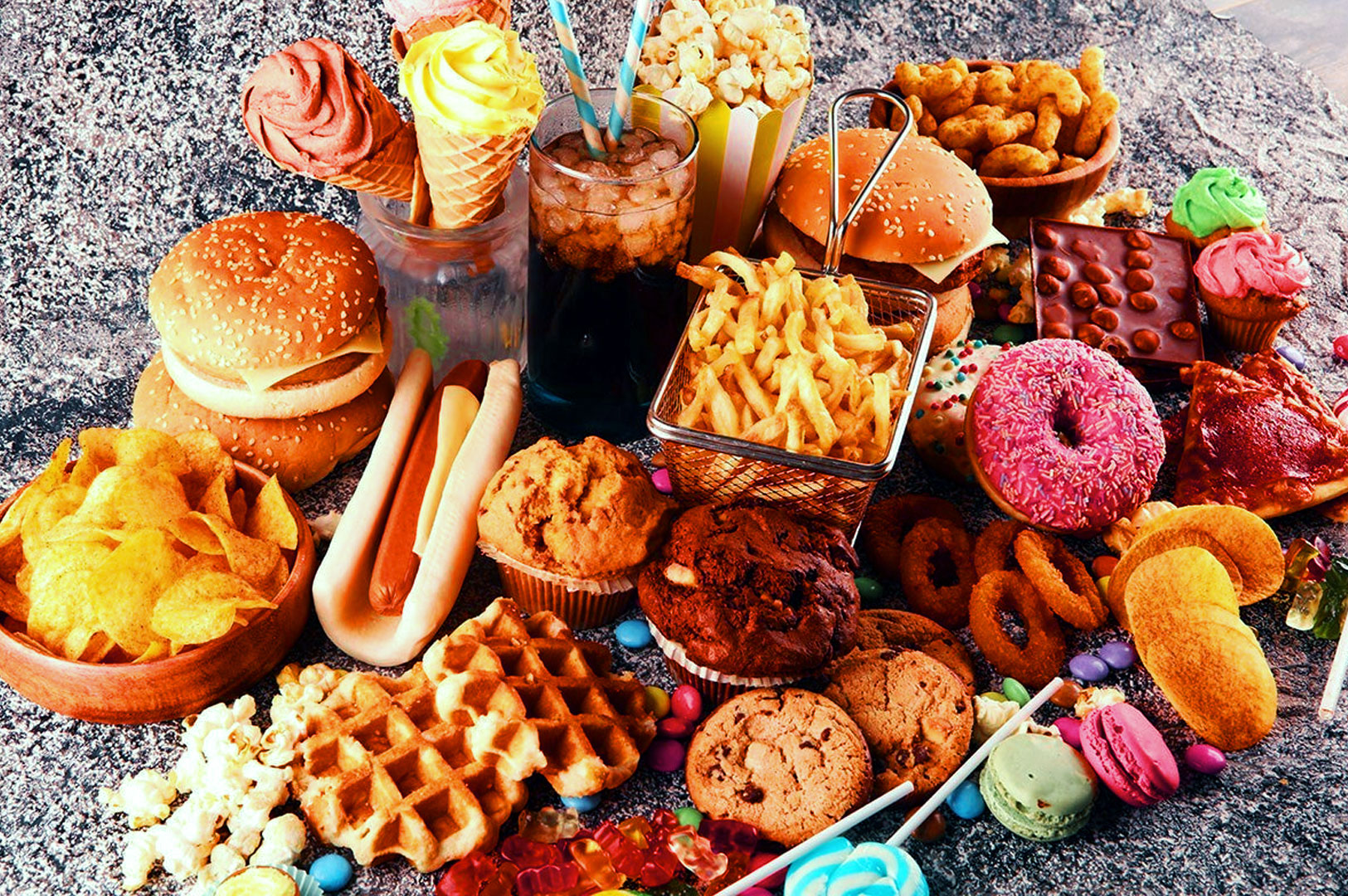 An assortment of junk food, including burgers, hot dogs, fries, donuts, cookies, waffles, candy, and popcorn, spread out on a textured gray surface.