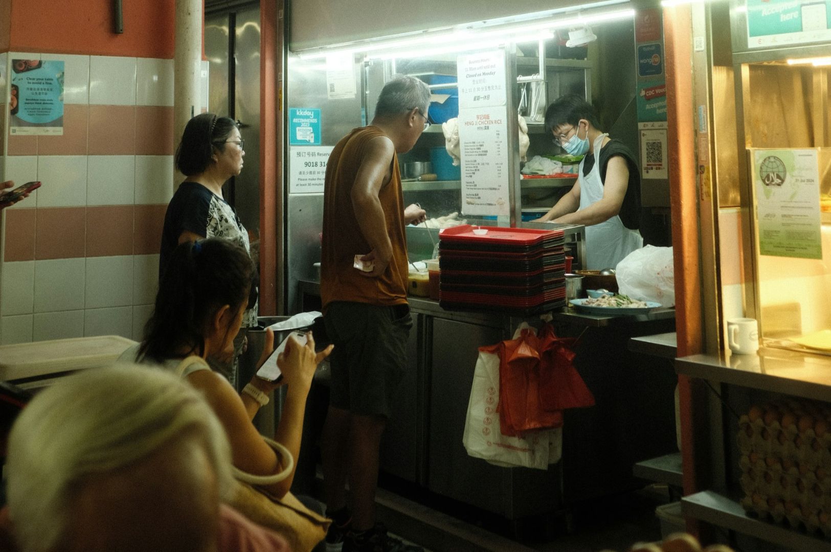 A small food stall scene with a man in an orange tank top ordering food. The vendor wears a mask and apron. Customers wait, creating a busy atmosphere.