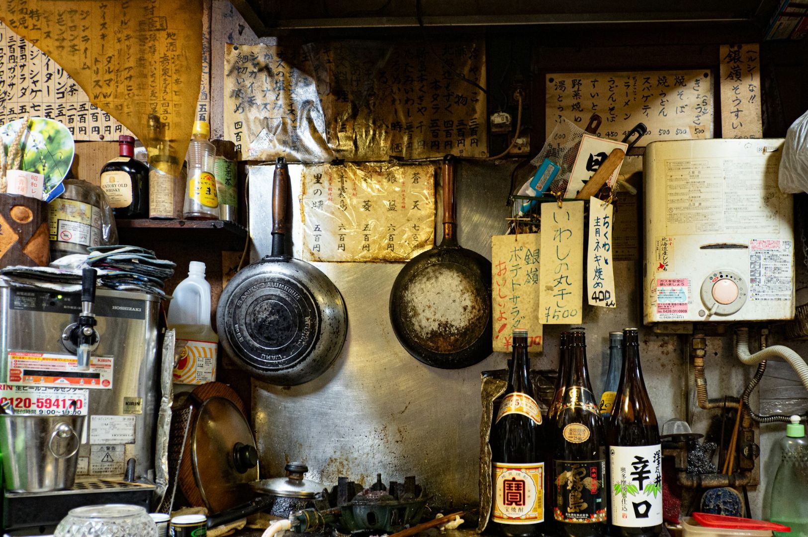 A cluttered, cozy Japanese kitchen with hanging pans, sake bottles, paper menus, and vintage decor. The warm, rustic setting evokes a nostalgic atmosphere.