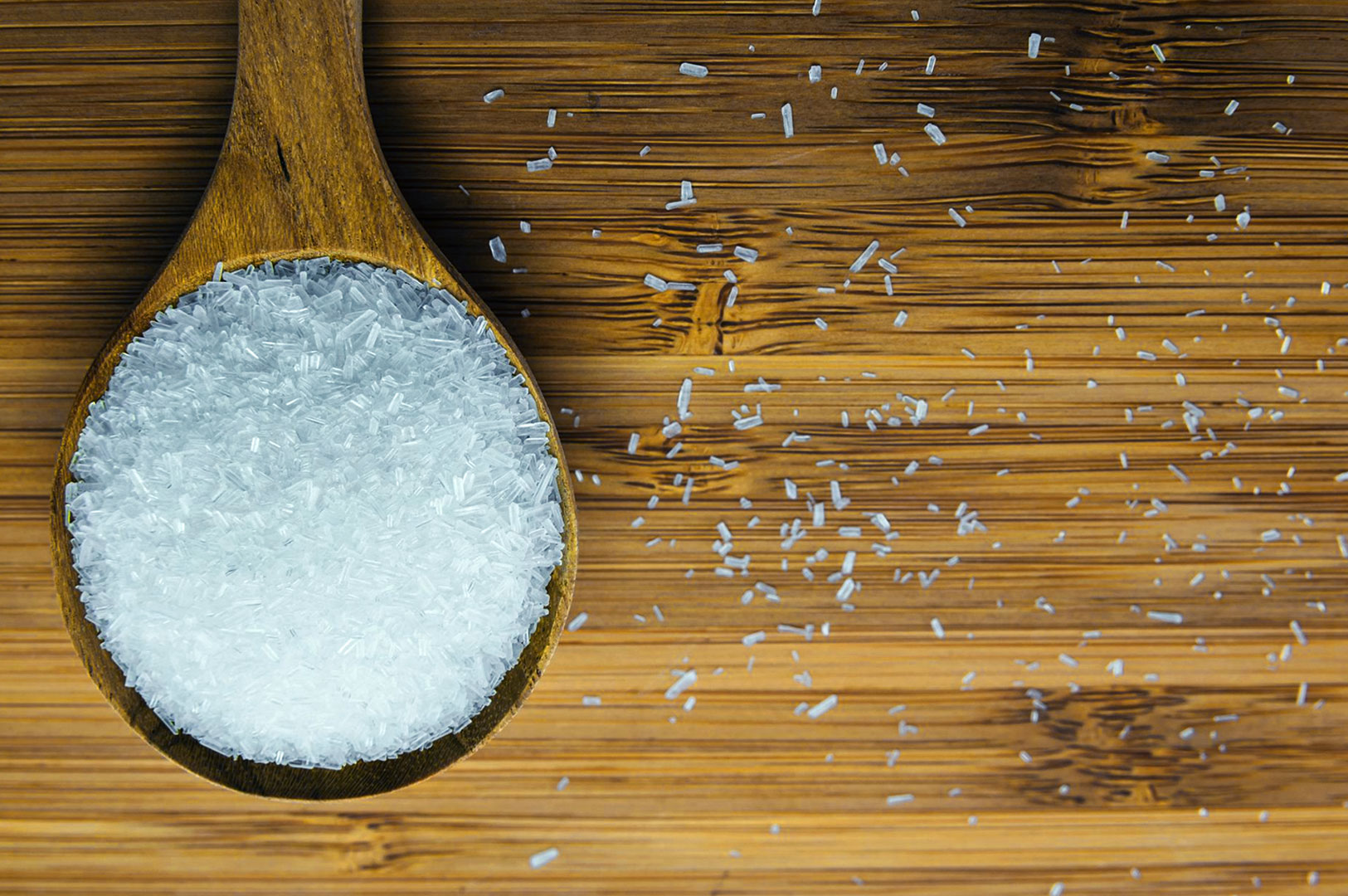 Wooden spoon filled with white crystalline powder, likely salt or MSG, on a bamboo surface. Some crystals are scattered around, creating a textured pattern.