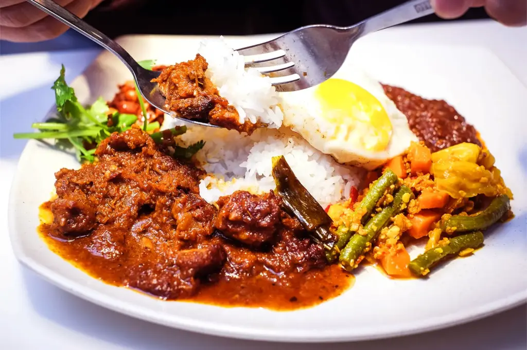 A plate of nasi lemak featuring beef rendang, white rice, a sunny-side-up egg, sambal, and mixed vegetables, evoking a vibrant and appetizing presentation.