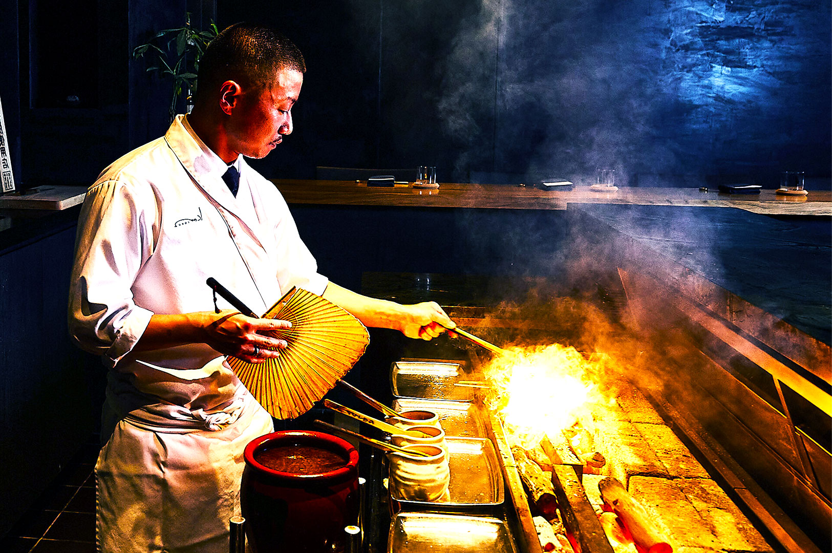 Chef in a white uniform skillfully fans flames on a grill, creating a vibrant, intense atmosphere. The dimly lit kitchen enhances focus and action.