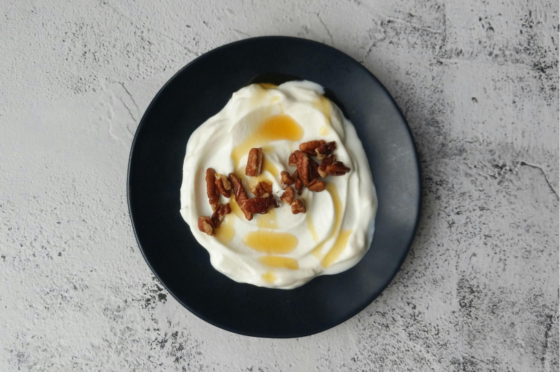 A top-down view of thick Greek yogurt on a matte black plate, topped with pecan pieces and a drizzle of honey.