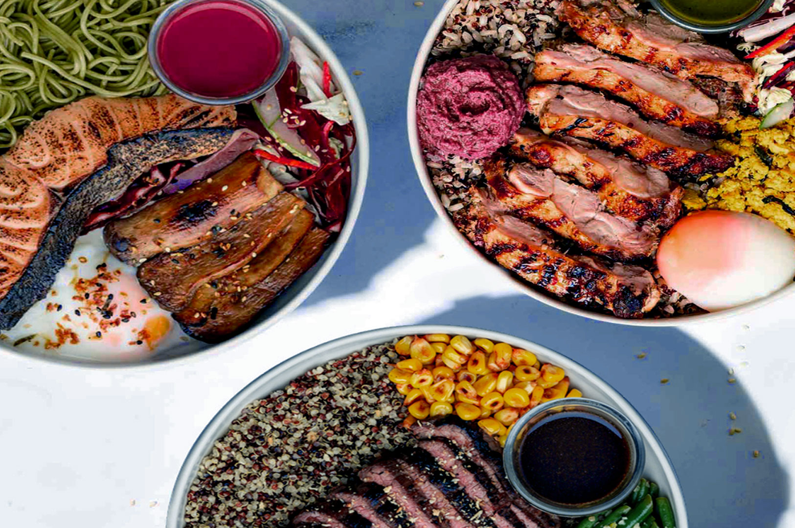 Three colorful bowls of food on a table. One has salmon and green noodles, another with grilled chicken and grains, the last with beef, quinoa, and corn.