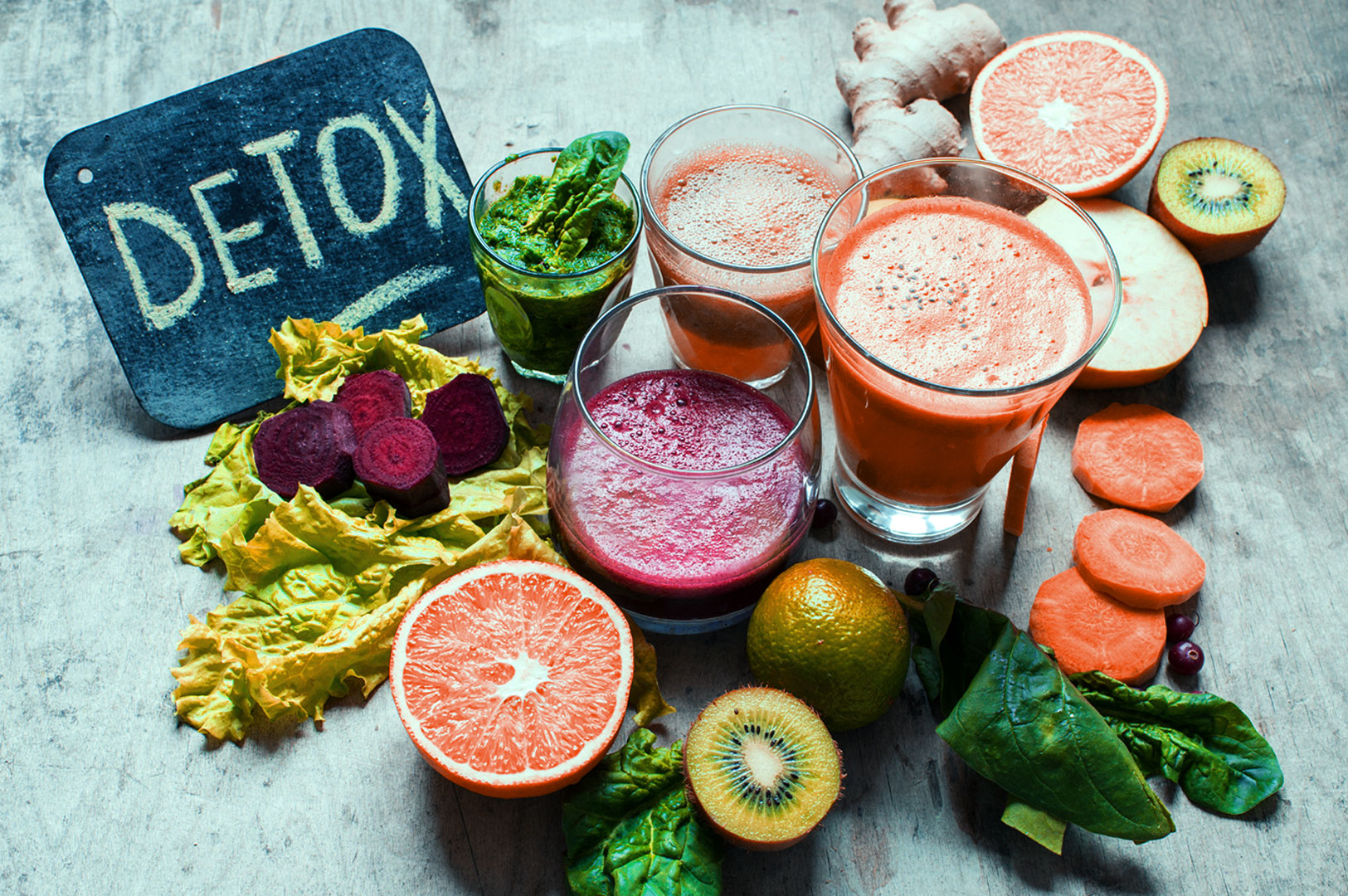 Three vibrant juice glasses surrounded by fresh fruits and greens, including grapefruit and kiwi. A chalkboard reads "Detox," suggesting health and vitality.