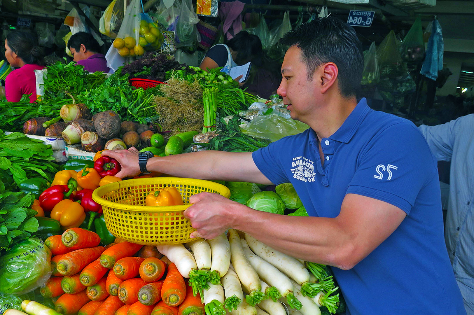 A person in a blue shirt selects red and yellow bell peppers from a vibrant vegetable market stand, surrounded by carrots, radishes, leafy greens, and other produce. The atmosphere is lively and bustling.