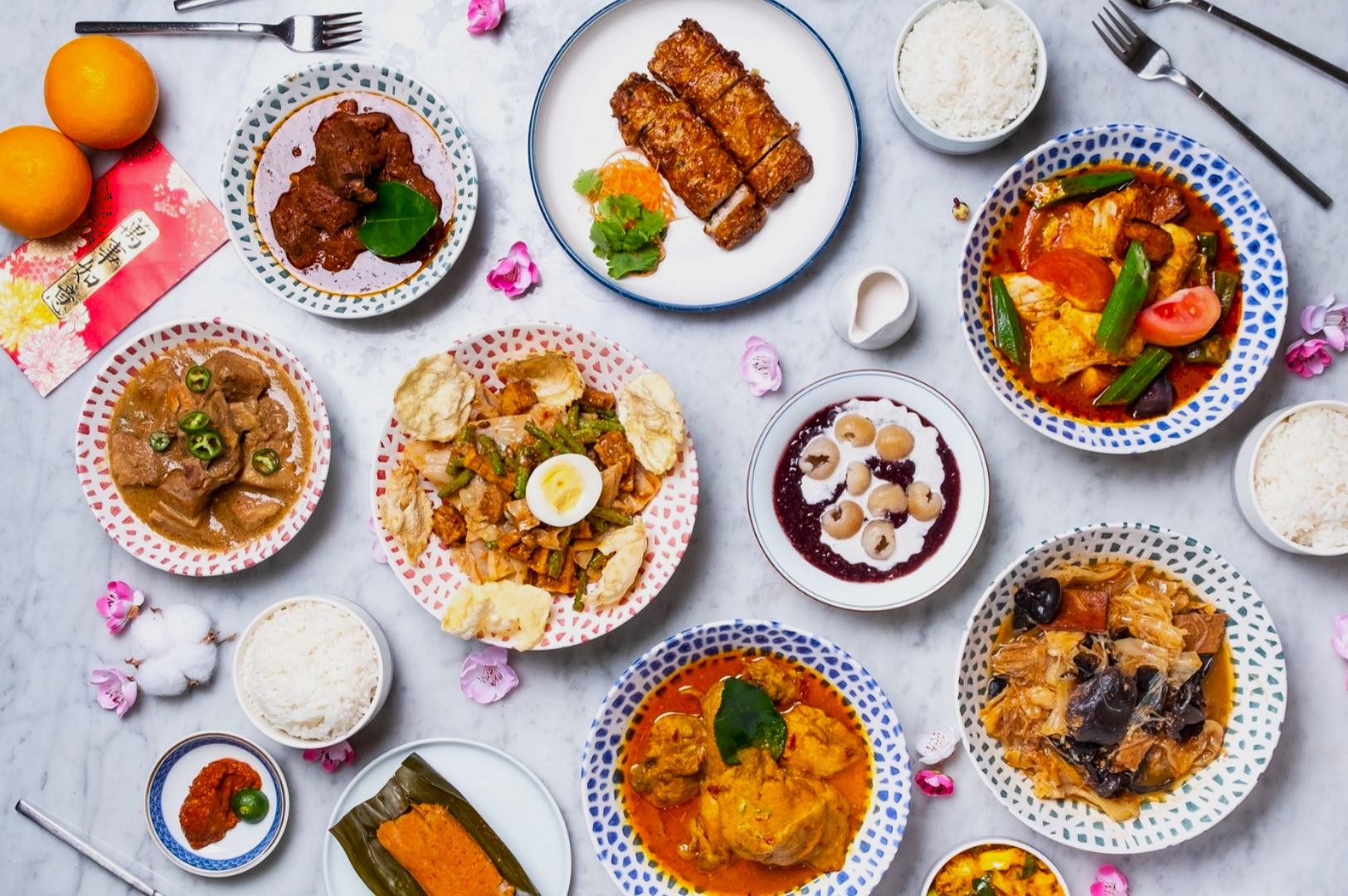 An array of colorful Asian dishes on a marble table, including meats, rice, and vegetables. Bright oranges and pink flowers add a festive touch.