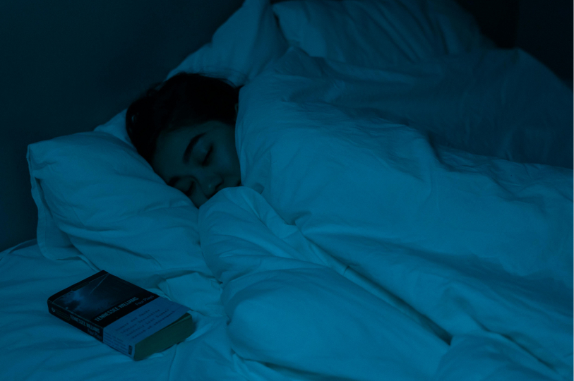 A person sleeping soundly under white covers in a dim, blue-lit room, with a book resting on the bed beside them.