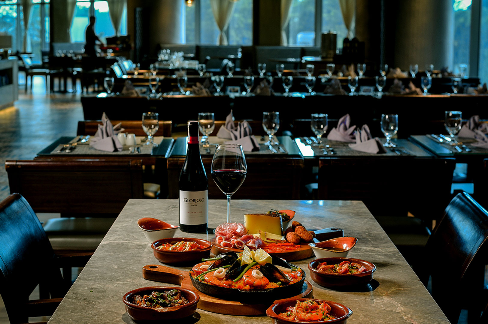 A lavish table setting in a dimly lit restaurant features Spanish tapas, a bottle of red wine, and a filled wine glass, evoking a cozy, elegant dining atmosphere.