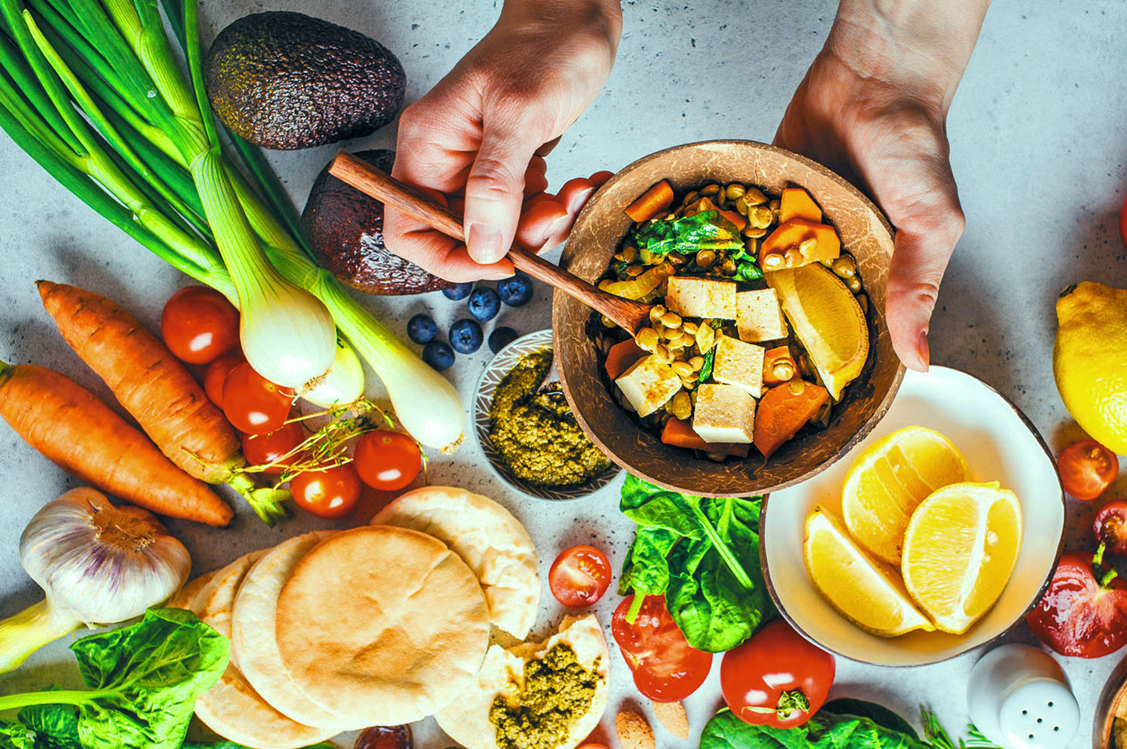 Hands holding a bowl of vibrant salad with tofu, corn, and lemon wedges above a table scattered with fresh vegetables and pita bread, conveying healthy dining.